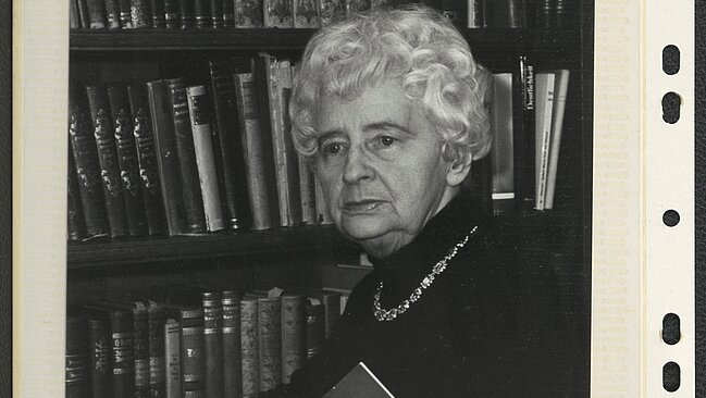 Schwarz-weißes Foto von Person die mit einem Buch vor einem Bücherregal posiert.